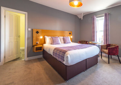 Photograph of room 6 looking towards bed with door leading to en-suite showeroom, purple tartan soft furnishings and occasional chairs with table