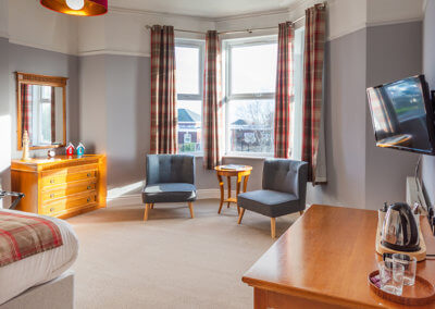 Photograph of the room 3 looking towards the bay window and seating area with two comfy grey chairs, occasional table and wooden chest of draws to the side.